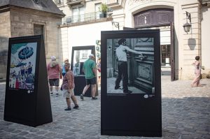 People walk past outdoor photo displays on a cobbled street in Paris, with buildings and an arched doorway in the background, evoking the charm of 1970. Some people are looking at the photos whilst others are walking by.