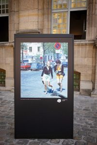 A display board stands on a cobbled street, featuring a photo of two women walking on a sunny day with cars and trees in the background, capturing the timeless charm of Paris 1970.