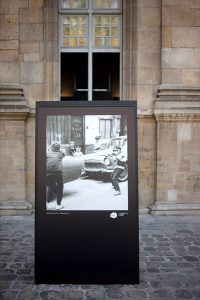 A black-and-white photo of children playing near a car is displayed on a black stand outside, in front of a stone building with a window above, capturing the timeless spirit of 1970s Paris.