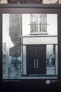 Black and white photo of a "CAFE BAR" entrance in 1970s Paris; a man is seen through the doorway, standing outside and holding a mobile. To the left is a worn wall. The image is part of a display panel.