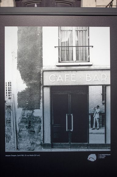 Black and white photo of a "CAFE BAR" entrance in 1970s Paris; a man is seen through the doorway, standing outside and holding a mobile. To the left is a worn wall. The image is part of a display panel.