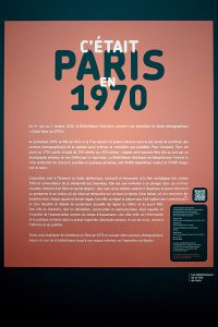 A sign with French text introduces a Paris 1970 photo exhibition at the Bibliothèque historique. It has a large title, event details, description, and a QR code. The background is red with black and white text.