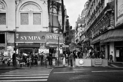 Black-and-white photo of a busy street with people queued outside a “SYMPA” shop, some carrying bags. Buildings surround the area, and a “Route Barrée” sign blocks part of the street, evoking scenes typical during periods of Renovation in Paris. In the background, Montmartre Basilica can be seen rising above the cityscape.