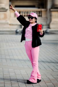 A woman, Elma Bailleul, dressed in pink trousers, a pink shirt, and a pink cap poses outdoors, holding a red mobile and pointing upwards with one arm. She is also wearing a black jacket and sunglasses.