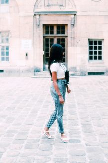Vivienne Jamet, a woman with long hair, wearing a white top, checked trousers, and sandals, walks on a cobbled courtyard in front of an old building with arched doors and windows.