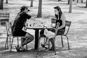 Two people, Maurice and Alice, sit at an outdoor table in a park, playing chess. Maurice is moving a piece while Alice watches attentively. Both appear focused. Trees and empty chairs are in the background. The image is in black and white.