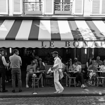Ali Akbar, dernier vendeur de journaux à la criée parisien A street performer in white walks past people sitting at outdoor tables under a striped awning at a café called “Le Bonaparte.” The scene is lively and relaxed, with patrons watchingA li Akbar, the last Parisian newspaper vendor hawking his wares.