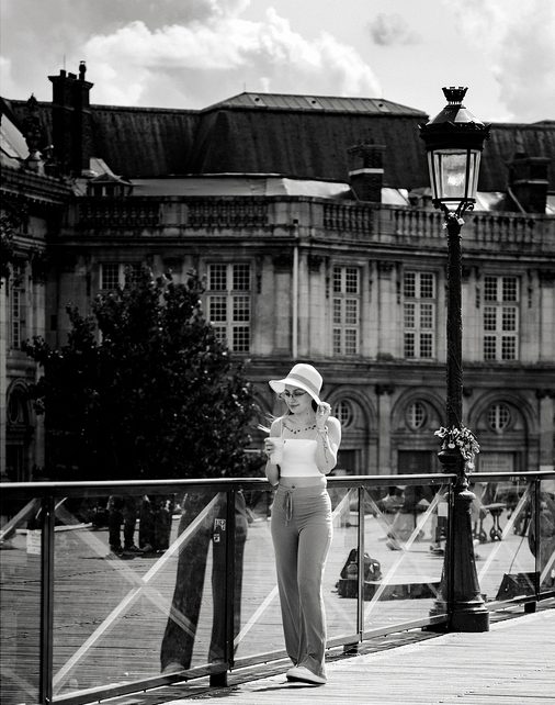 A woman in a hat and sunglasses walks on a bridge, holding her mobile and smiling. An ornate building and a streetlamp are in the background. The image is black and white, capturing Sally Leloup’s effortless style as she enjoys the city scene.