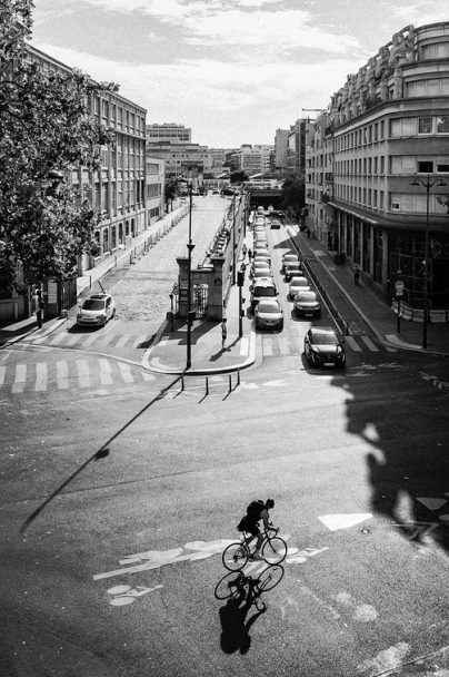 L'ombre du vélo A cyclist rides through a sunny city junction on their vélo, casting an ombre on the road. Cars queue along the street, surrounded by tall buildings and trees on a clear day.