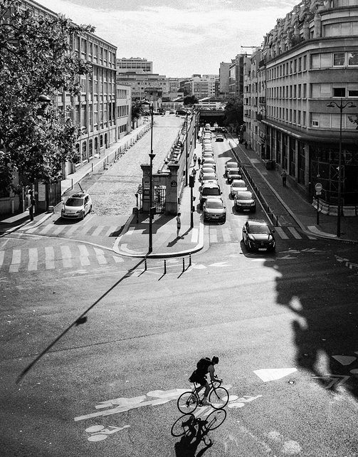 A cyclist rides through a sunny city junction on their vélo, casting an ombre on the road. Cars queue along the street, surrounded by tall buildings and trees on a clear day.