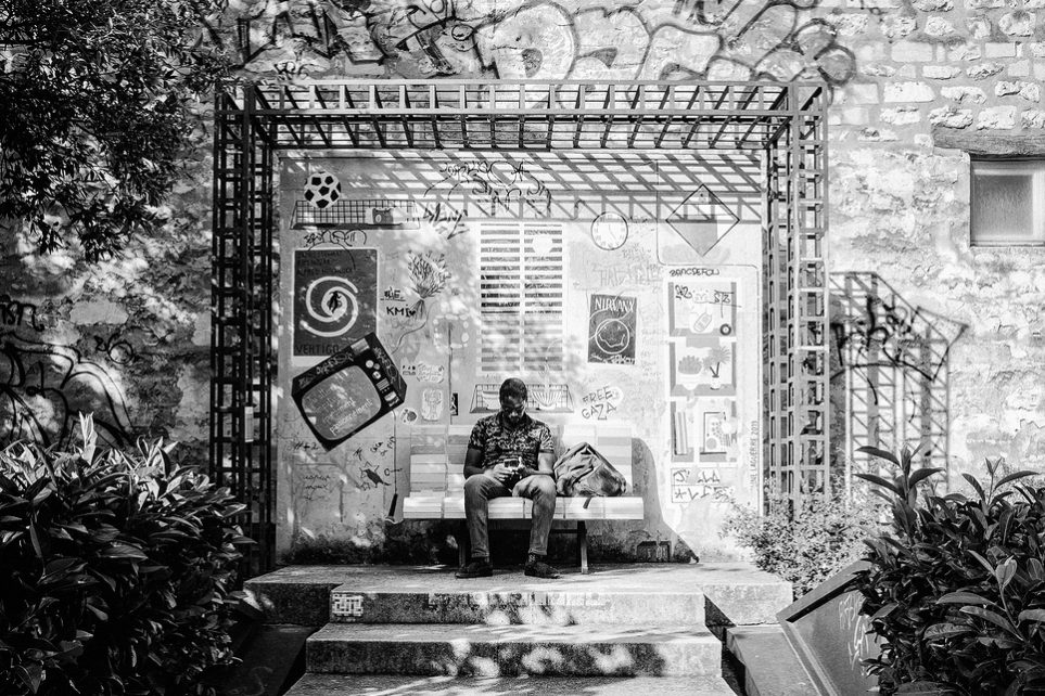 A man sits on a bench under a metal trellis, surrounded by plants and graffiti art on a stone wall, reading a book in bright sunlight. The scene is in black and white, evoking the quiet introspection often found in Thomas Chopin’s works.