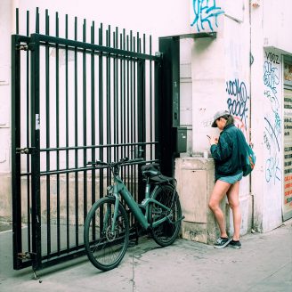 Frankie Guillemin, in a cap and jacket, stands by a graffiti-covered wall, using their mobile. A bicycle is parked against a black metal gate nearby.