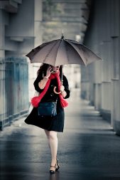 A woman in a black coat and red scarf walks under a brolly on a rainy day, talking on her mobile in a city corridor—a scene reminiscent of a modern Josephine Beauvais, graceful yet purposeful amidst the urban rain.