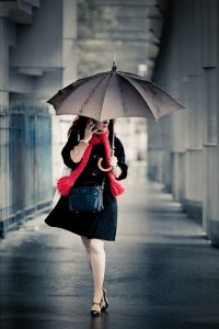 A woman in a black coat and red scarf walks under a brolly on a rainy day, talking on her mobile in a city corridor—a scene reminiscent of a modern Josephine Beauvais, graceful yet purposeful amidst the urban rain.