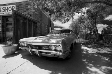 A vintage car is parked in front of a small brick shop on a sunny day, with trees casting shadows over the scene and a large planter visible near the doorway.