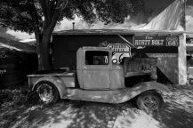 An old, rusted pickup lorry sits by a building with a “Historic Route 66” mural and “The Rusty Bolt” sign in Seligman, Arizona. A large tree shades the left side of the scene.