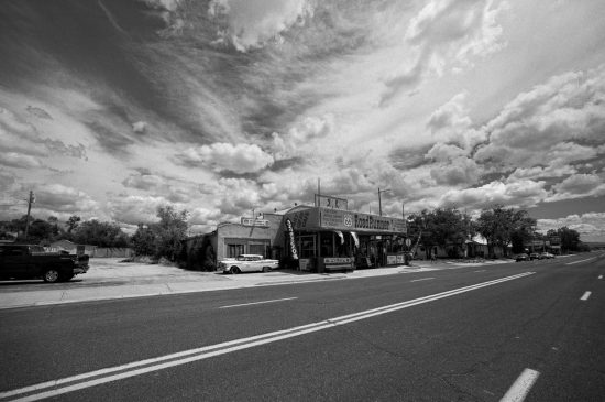 A wide road passes by an old-fashioned roadside market with vintage cars parked outside. The sky above is dramatic, filled with swirling clouds.