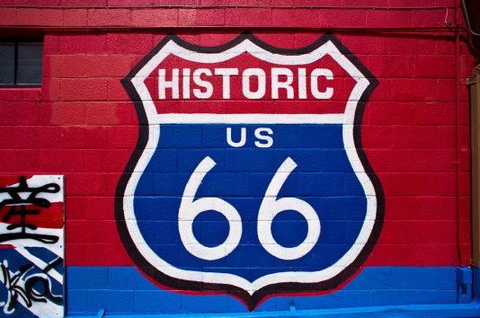 A mural of the Historic US Route 66 shield is painted on a red brick wall with blue and white accents in Seligman, Arizona. Part of another mural with black symbols is visible on the left.