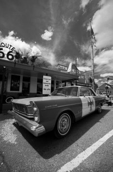A vintage car parked outside a Route 66 diner under a partly cloudy sky, with an American flag and classic car signs visible in the background. Black and white photo.