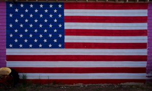 A large American flag is painted on the side of a wooden building with horizontal boards in Seligman, a famous stop along Route 66 in Arizona. There’s a small yellow object and some grass at the bottom left corner.