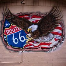Mural of a bald eagle flying in front of a waving American flag and a Route 66 road sign, painted on a brick wall in Seligman, Arizona.