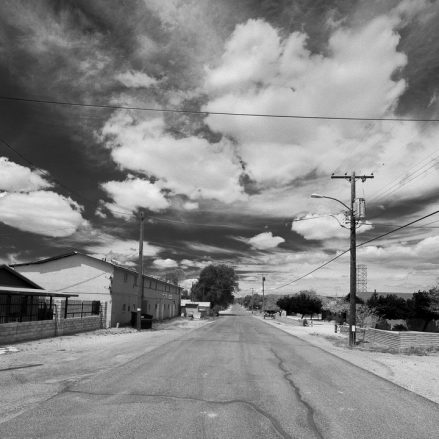 Black and white photo of an empty street in a residential area, with houses, power lines, and dramatic clouds filling the sky.