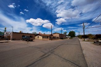 A wide, empty street in a small town with a few buildings, a parked pickup lorry, power lines, and a bright blue sky with scattered clouds.