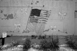 A faded mural of an American flag and four bows is painted on a worn, peeling wall. Overgrown weeds line the ground beneath the mural.