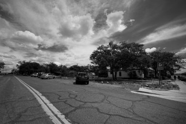 A cracked road leads past parked cars and houses, with large trees and a dramatic, cloudy sky overhead in black and white.