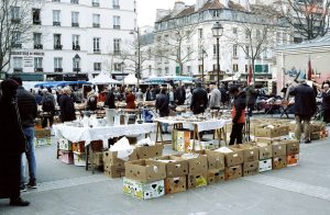 Outdoor flea market scene with people browsing tables of various goods, reminiscent of a lively Paris market. Cardboard boxes filled with items are stacked in the foreground, while blocks of flats and leafless trees are visible in the background.