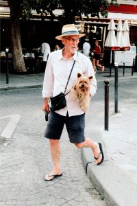Older man Damien Schneider, in a straw hat, glasses, pink shirt, shorts, and flip-flops walks on a city street holding a small brown dog. There are people, umbrellas, and trees in the background.