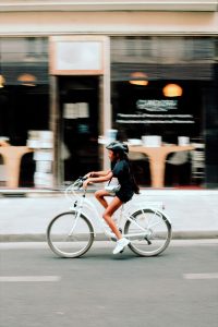 A person wearing a helmet rides a white bicycle along a city street, with a blurred café and tables in the background.