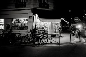 A black and white photo of a corner bakery at night, illuminated from within. Several bicycles are parked by a metal fence outside, and nearby streets are mostly empty.