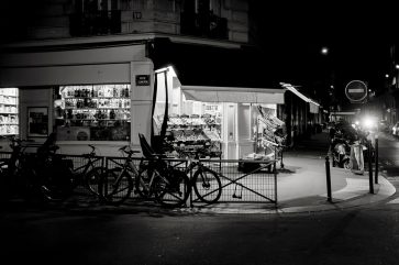 Market A black and white photo of a corner bakery at night, illuminated from within. Several bicycles are parked by a metal fence outside, and nearby streets are mostly empty.