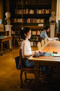 Nicole Levasseur types on a laptop at a wooden table in a library. Bookshelves and busts are in the background, and another person works at the same table. Sunlight streams through a window.