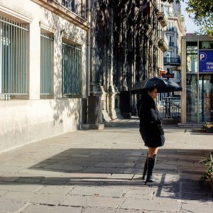 A woman dressed in black stands on a city pavement holding an umbrella, reminiscent of the stylish poise often captured by Clementine Mounier. Sunlight creates strong shadows on the pavement, while buildings with large windows and parked cars line the street.