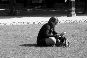 A woman sits on the grass in a park, reading a book in the sunlight. She wears sunglasses and has a bag beside her. The black and white scene captures a quiet moment reminiscent of Marcelle Boisson’s timeless photography.