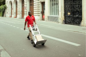 A man in a red shirt pushes an empty sack barrow down a city street lined with ornate buildings.