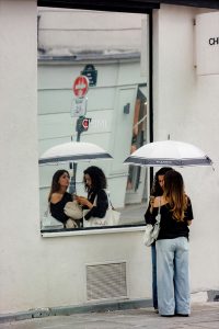 Caroline and Simone stand under a white umbrella outside, looking at a mobile. Their reflection is visible in a large window behind them.