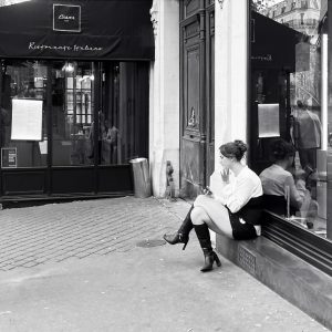 A woman, perhaps Angelique Baron, sits on a ledge outside a restaurant, looking at her mobile. She wears a white blouse, black skirt, and heeled boots. The scene is in black and white.