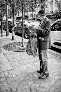 An older man in a hat and striped jacket stands on a city pavement reading a newspaper, holding a patterned bag and walking stick, with parked cars and trees nearby. Reminiscent of the candid street photography of Alain Bouyer, this black and white image captures a timeless urban moment.