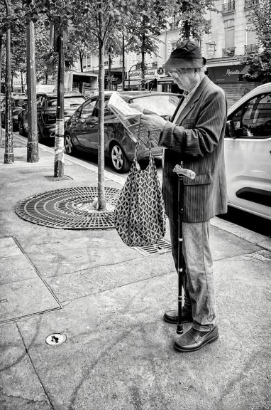 An older man in a hat and striped jacket stands on a city pavement reading a newspaper, holding a patterned bag and walking stick, with parked cars and trees nearby. Reminiscent of the candid street photography of Alain Bouyer, this black and white image captures a timeless urban moment.