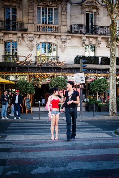 A couple walks across a zebra crossing in front of a Parisian café. Vivien, wearing a red top and shorts, chats and smiles with Casimir, who is dressed in black. The street and café are lively, adding to the vibrant atmosphere as they stroll together.