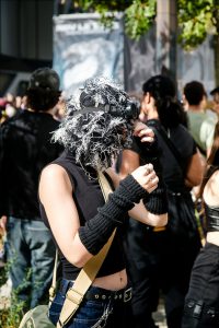 A person wearing a textured, shaggy mask with goggles, fingerless gloves, and a sleeveless top stands in a crowd at an outdoor event, adjusting their mask. Trees and people are visible in the background, capturing the vibrant energy of the 2026 Techno Parade in Paris.