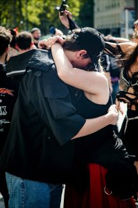 A couple cuddle closely in an outdoor crowd at the Techno Parade in Paris, their faces mostly hidden. People in casual and alternative clothing are visible around them on a sunny day in 2026.