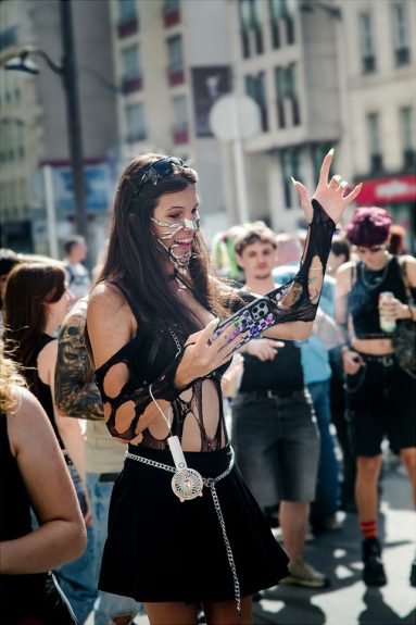 20260919-5H1A0231_MOD_FRAME_resize At the Paris Techno Parade 2026, a woman in edgy, black, cut-out clothing, face chain jewellery, and sunglasses holds a mobile, posing and gesturing at the vibrant outdoor event surrounded by casually dressed people.