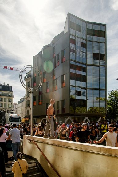 20260919-5H1A0236_MOD_FRAME_resize A crowd gathers in front of a modern glass building in Paris; a shirtless man stands on a ledge, and a metal sculpture is visible in the background under a partly cloudy sky—capturing the vibrant energy of the Techno Parade 2026.