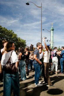 20260919-5H1A0238_MOD_FRAME_resize A group of people gathers outdoors near a tall monument under a blue sky during the Paris 2026 Techno Parade. Some are talking, others are dancing, and most appear relaxed and cheerful. Trees and streetlights are also visible in the background.