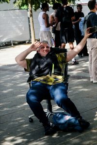 A man sitting on an office chair outdoors in Paris smiles and raises his arm for a selfie during the lively Techno Parade 2026. People stand and talk in the background as sunlight filters through the trees, capturing the vibrant energy of the event.