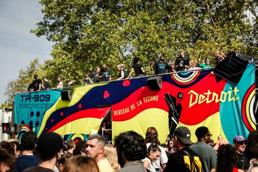 20260919-5H1A0245_MOD_FRAME_resize A colourful parade float with “Detroit” and “TR-909” written on it moves through a crowd during a sunny outdoor festival, reminiscent of the lively energy at the Paris Techno Parade. People dance and enjoy music on top of the float, while many watch from below, celebrating together in anticipation of 2026’s vibrant events.
